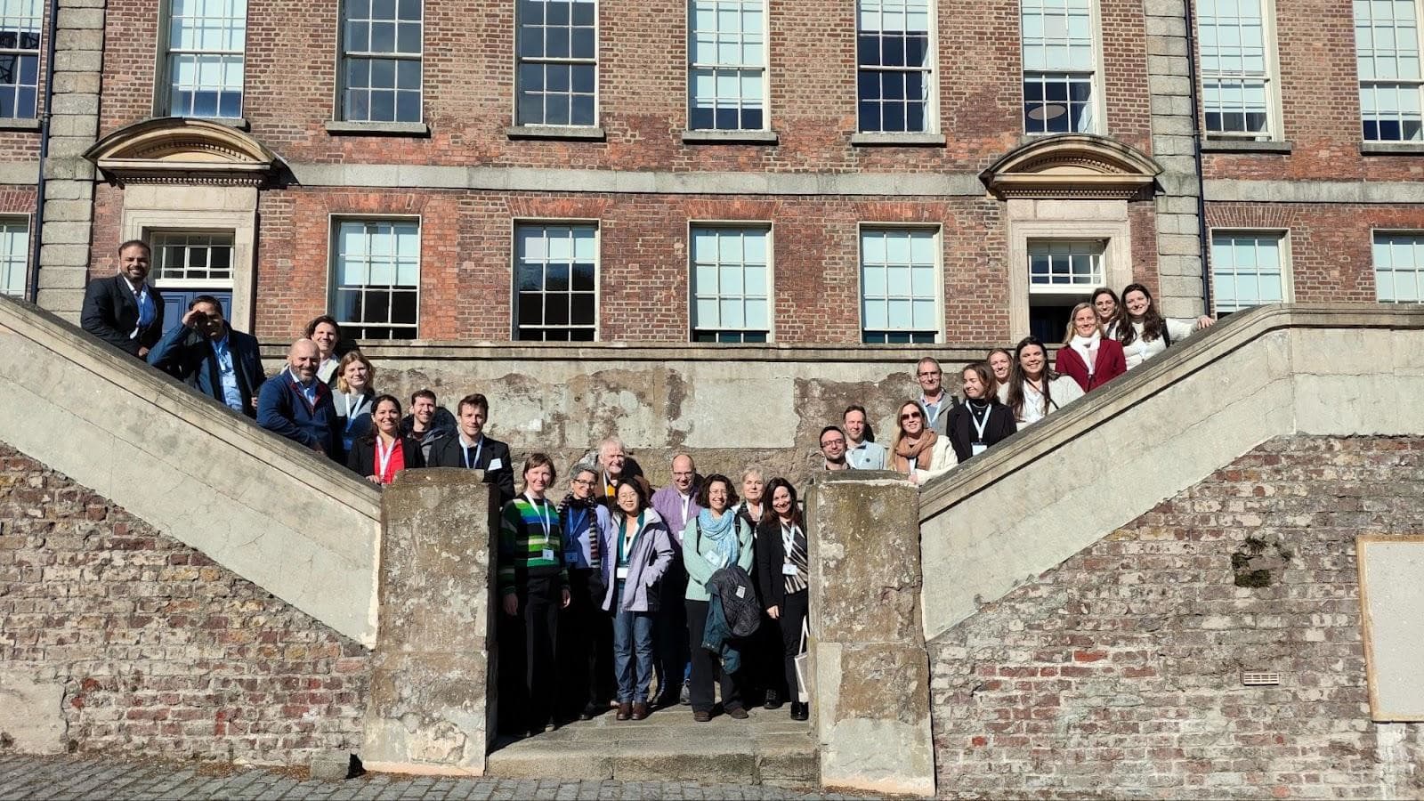 A diverse group of individuals stands on stone steps in front of a historic brick building with large windows.